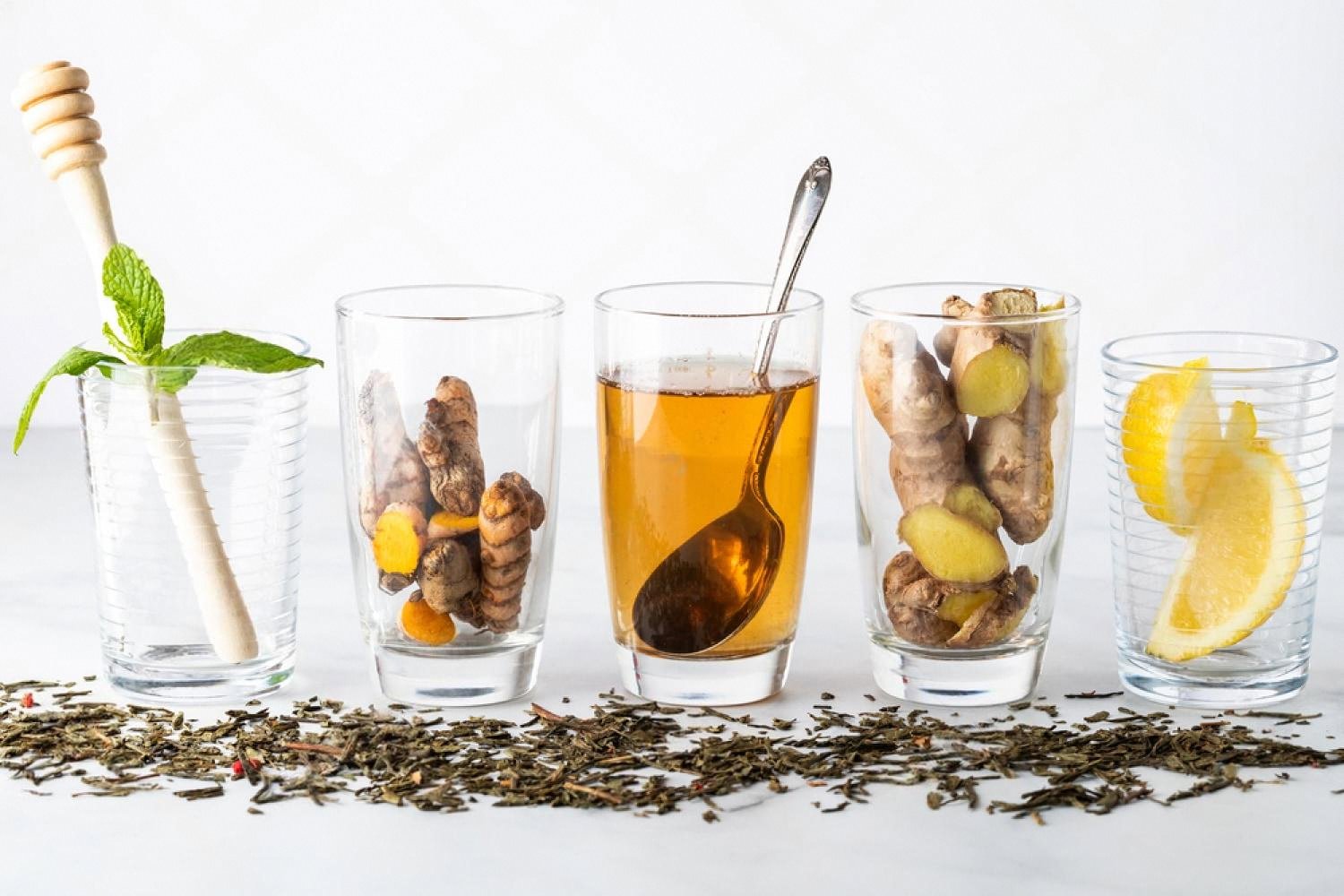 Row of glass tumblers with mint, turmeric, ginger, honey and lemon over scattered tea leaves